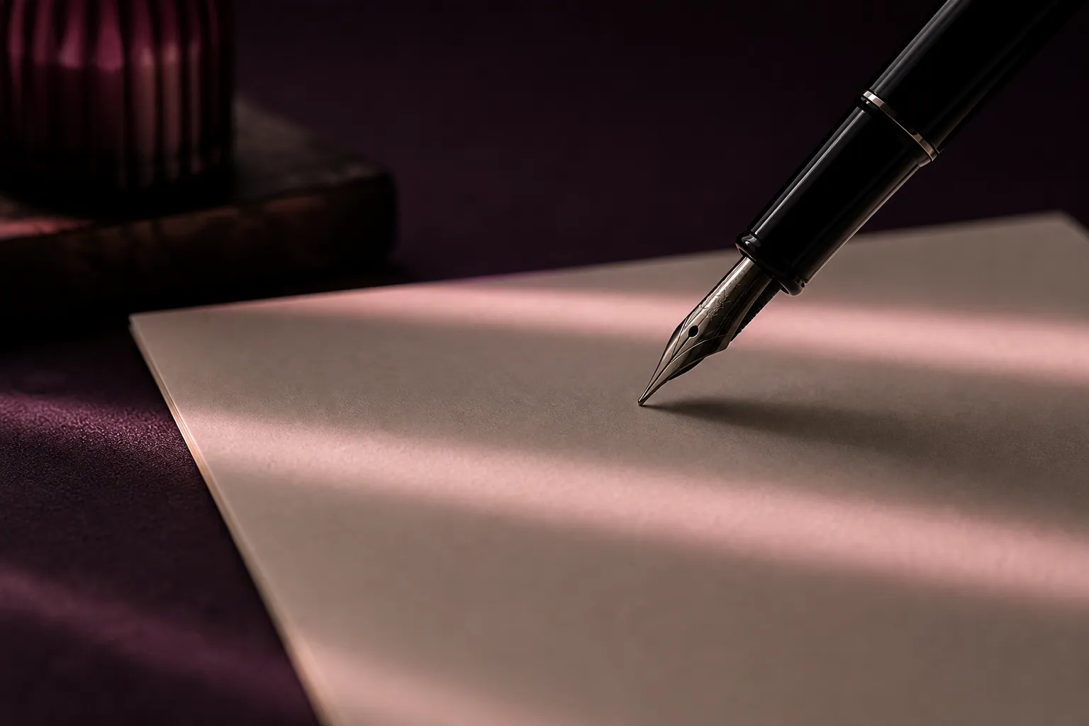 A black fountain pen hovering above blank cream paper crossed by soft bands of rose light.